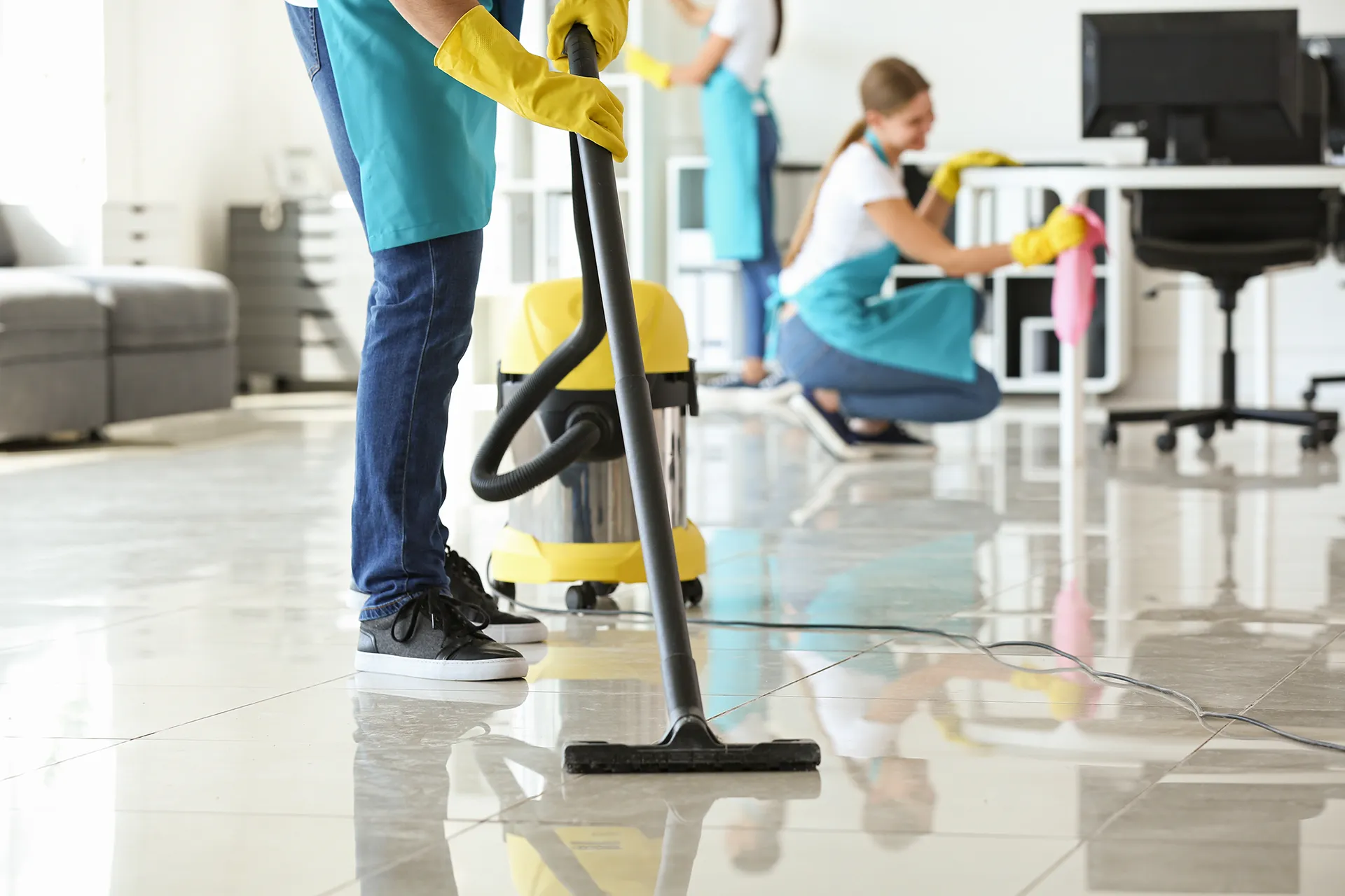 Professional office cleaners in turquoise aprons vacuuming tile floors and wiping down white desks in a bright, modern corporate workspace.