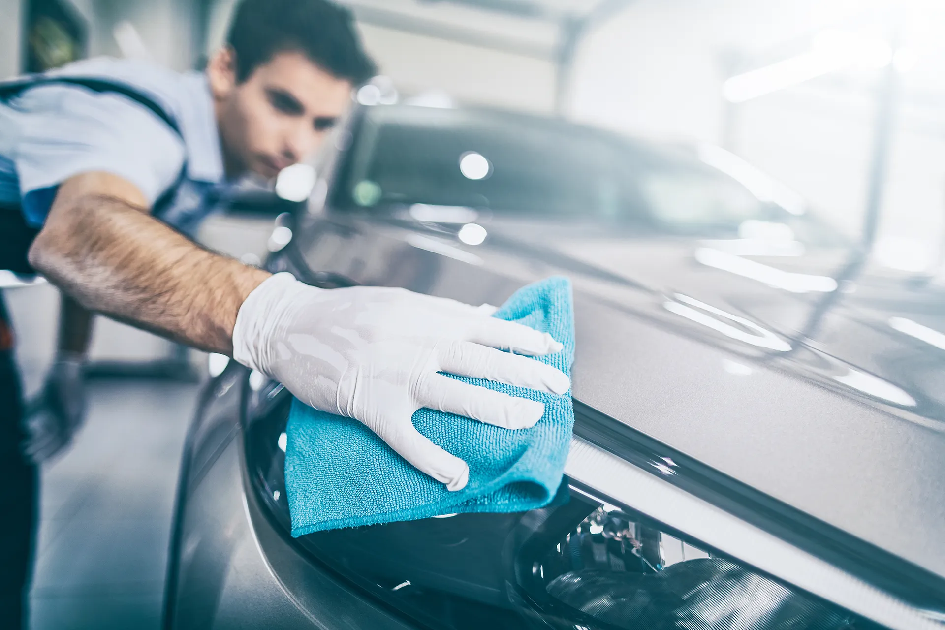 Professional automotive detailer wearing white protective gloves using a blue microfiber cloth to polish the exterior headlight and hood of a silver car.