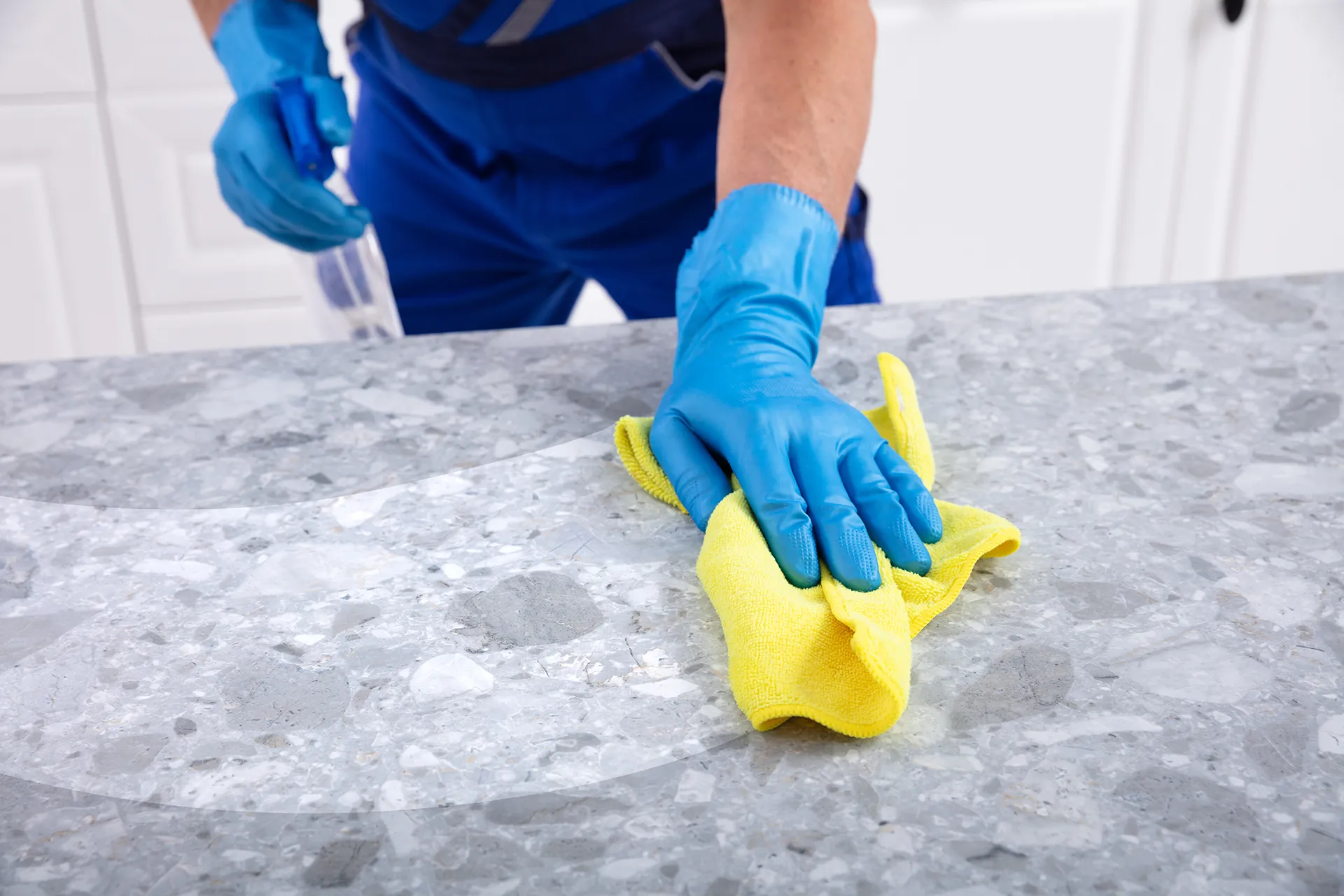 Close-up of a professional cleaner wearing blue nitrile gloves using a yellow microfiber cloth and spray bottle to sanitize a grey speckled granite countertop.