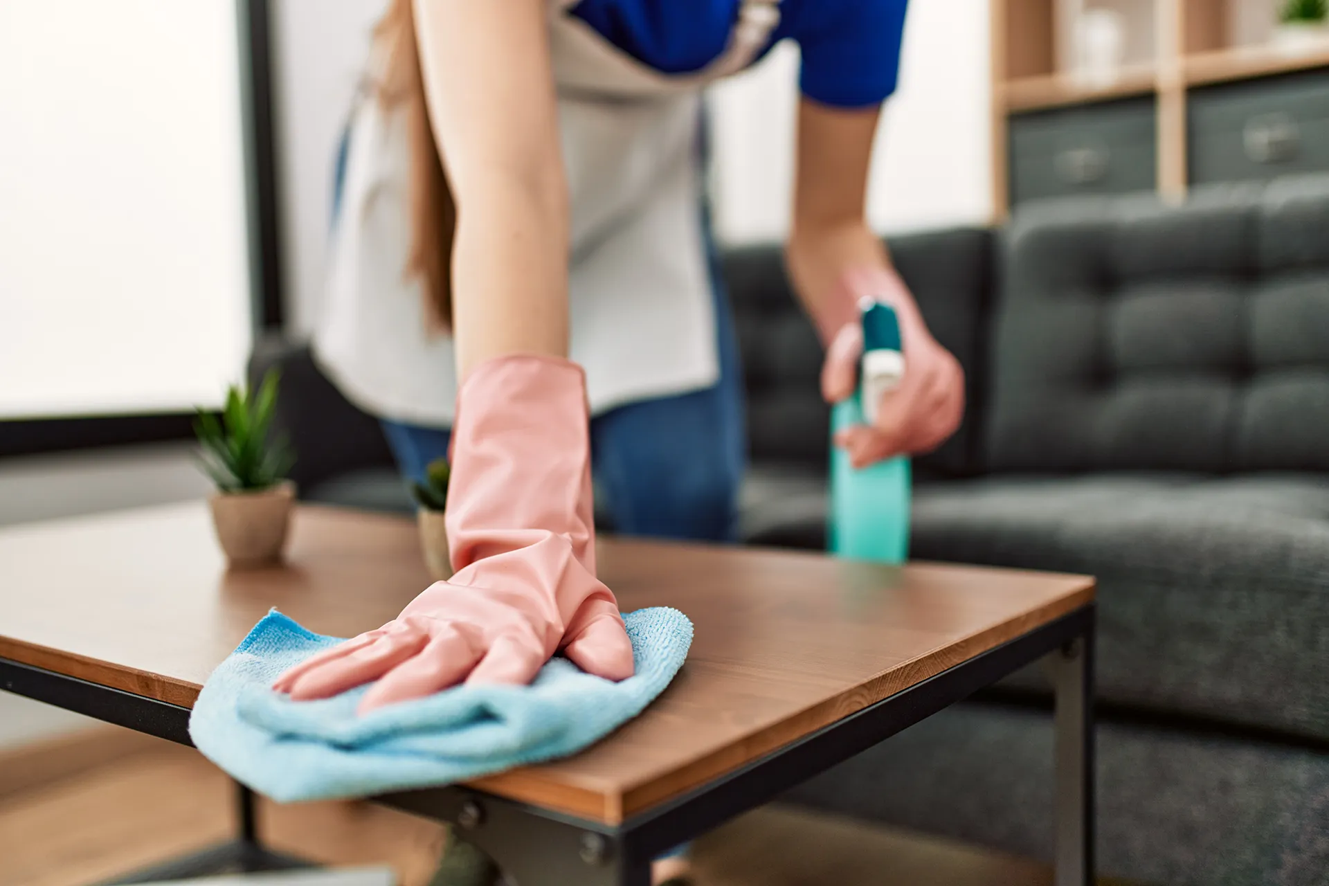 Professional house cleaner wearing pink rubber gloves using a blue microfiber cloth and spray bottle to wipe down a wooden coffee table in a modern living room.