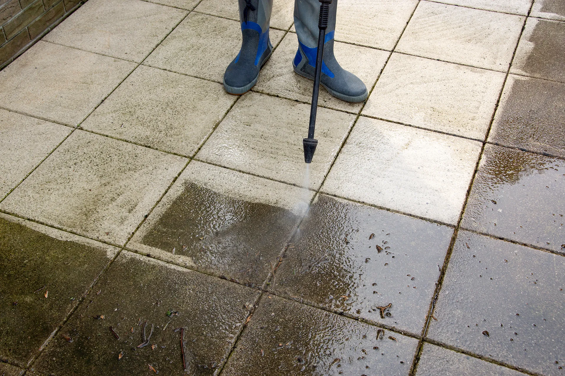 High-pressure water spray being used to deep clean outdoor stone patio tiles, removing dirt and grime to reveal the original light-colored surface.